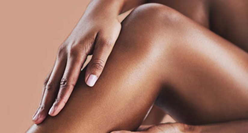 Close-up van een hand die zachtjes over een glad, gebruind been strijkt. De huid ziet er gehydrateerd uit en de achtergrond is een neutrale kleur.