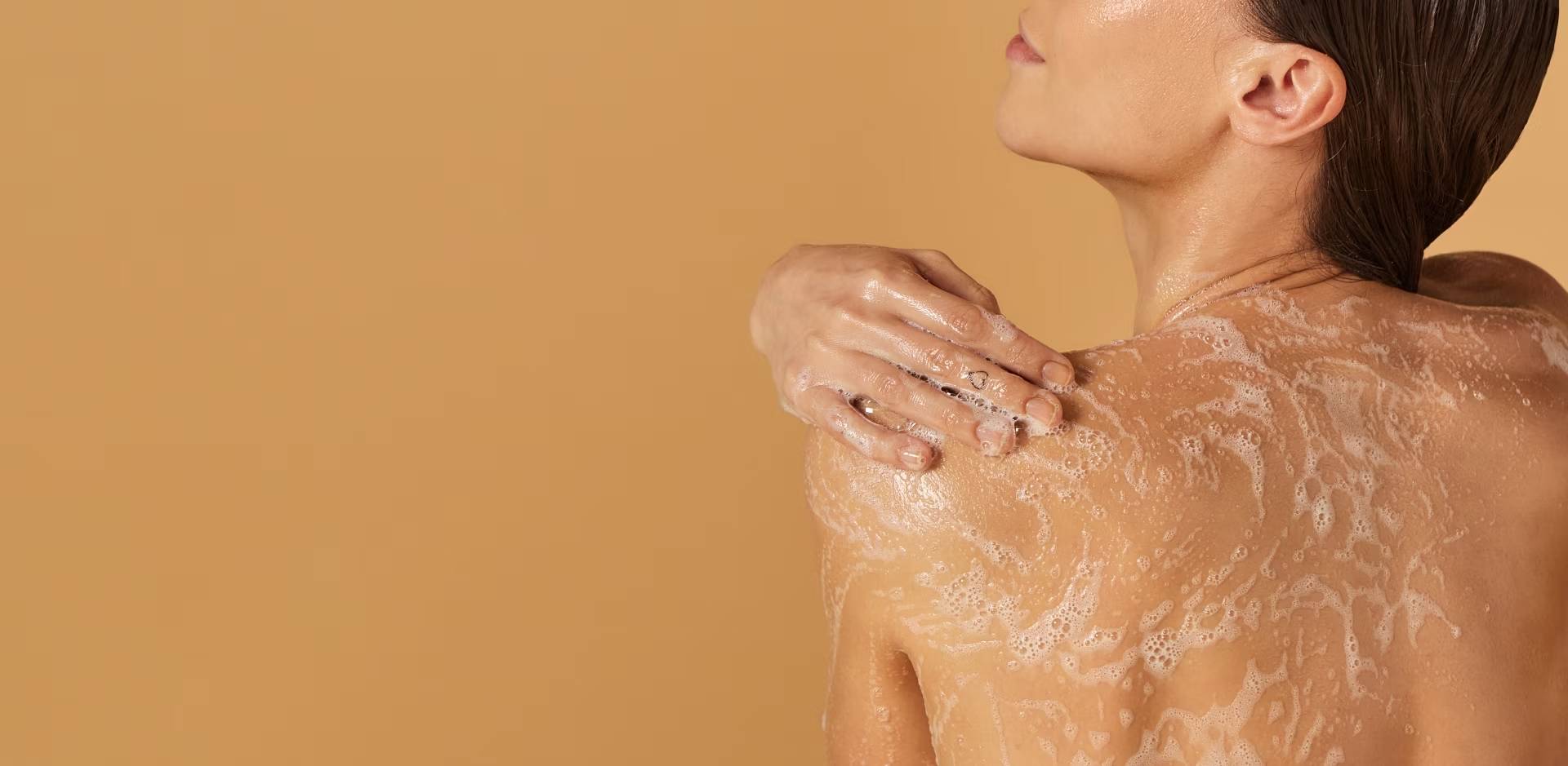 Een vrouw, gezien op haar rug, die haar schouder wast met een rijk schuim. Haar hand rust op de ingezeepte huid en de achtergrond is effen beige.