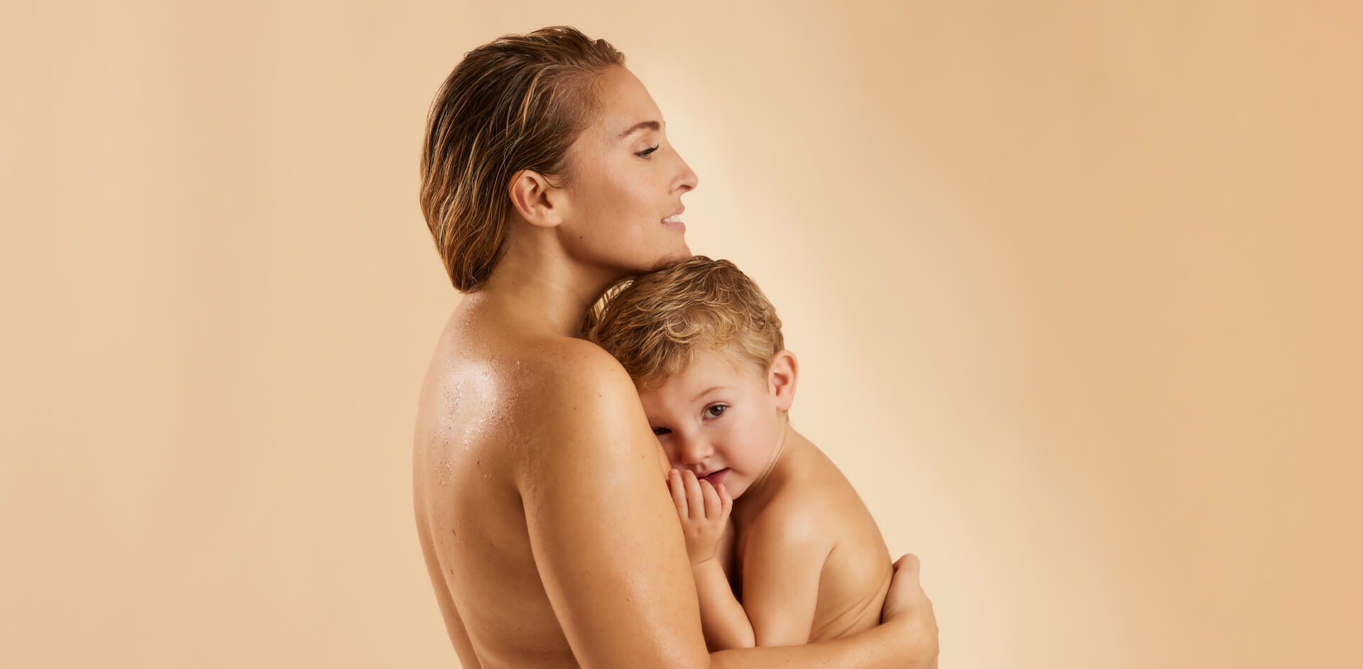 Een vrouw met nat haar houdt een klein kindje teder vast. Het kindje met blonde krullen kijkt vanaf haar schouder de camera in.