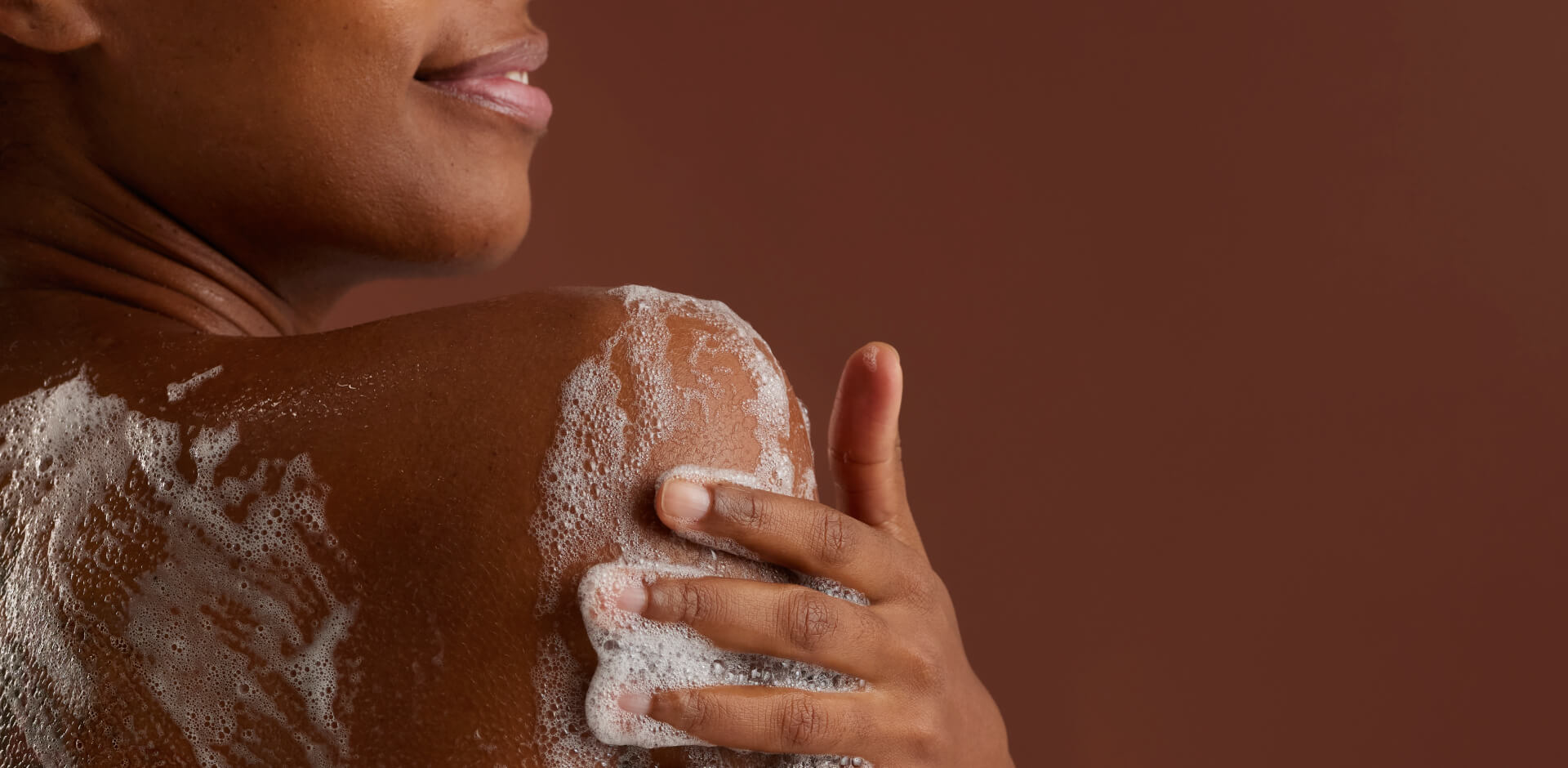 Close-up van de rug en schouder van een persoon met een donkere huid, die zichzelf inzeept met wit schuim tegen een bruine achtergrond.