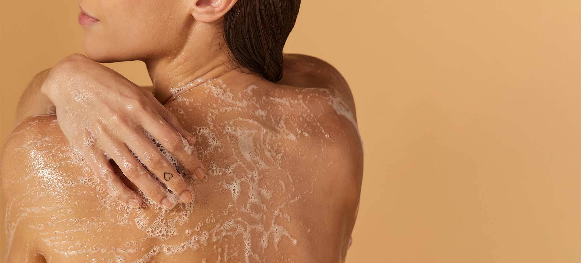 Een vrouw, gezien op haar rug, die haar schouder wast met een rijk schuim. Haar hand rust op de ingezeepte huid en de achtergrond is effen beige.