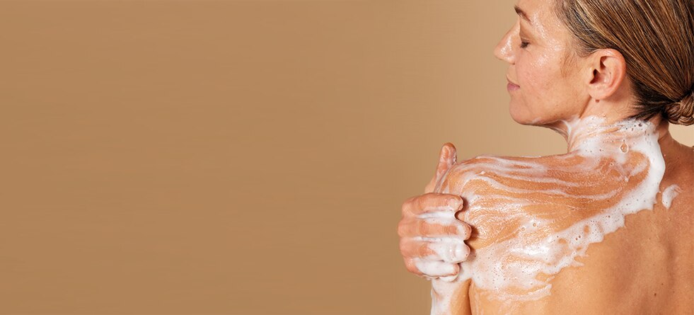 Een vrouw met een lichte huidskleur, gezien vanaf haar rug, die haar schouder inzeept met een rijk schuim tegen een beige achtergrond.