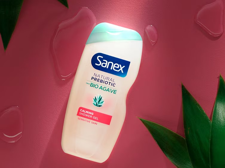 Sanex Natural Prebiotic Bio Agave Calmante Douchegel met roze label op een roze achtergrond met groene bladeren.