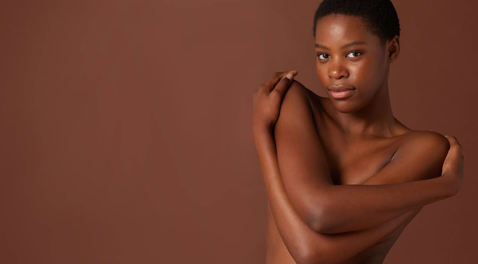 Een zwarte vrouw met kort haar omhelst zichzelf terwijl ze in de camera kijkt, tegen een bruine achtergrond.